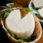 Formaggio primosale di capra artigianale in cestino di vimini con foglie aromatiche, su tavolo rustico in legno.