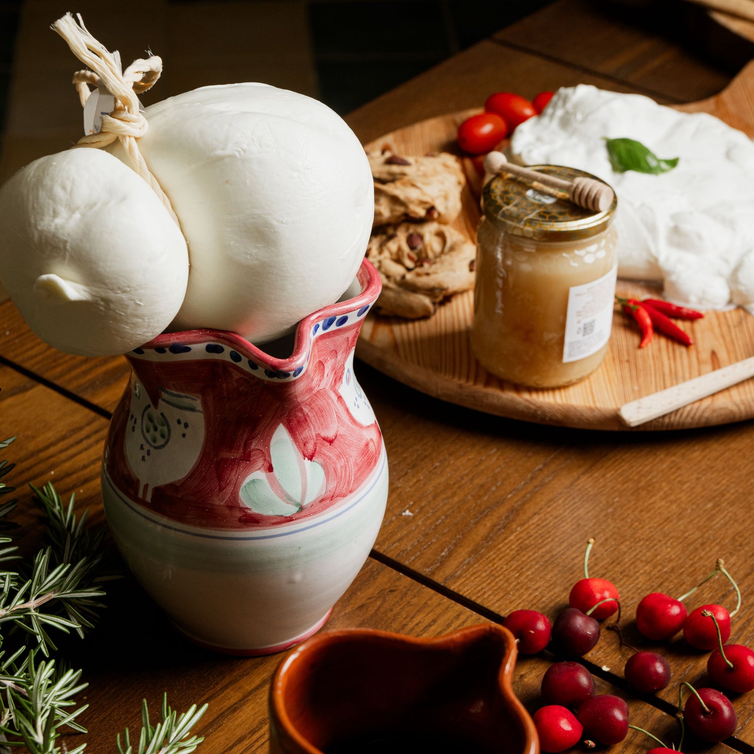 Caciocavallo artigianale legato con corda naturale, esposto in una brocca in ceramica decorata su tavolo rustico, con ciliegie, miele, pane e formaggi freschi sullo sfondo.