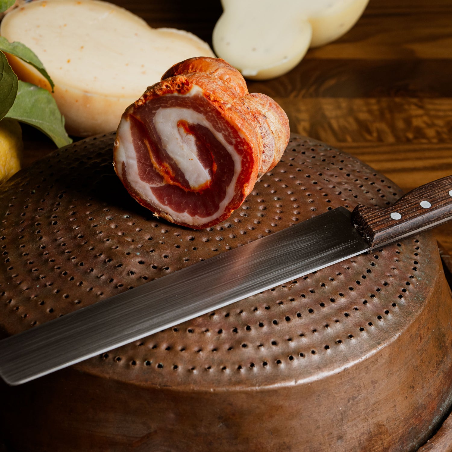 Pancetta arrotolata cilentana posizionata su un antico coperchio in rame, con coltello da cucina e ciliegie fresche sullo sfondo.