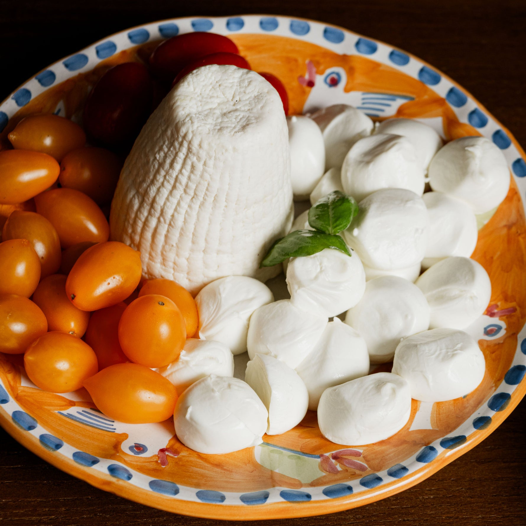 Ricotta dura al centro di un piatto decorato, circondata da pomodorini gialli, bocconcini di mozzarella e foglie di basilico, su tavolo in legno scuro con grappolo d’uva verde in primo piano.