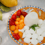 Ricotta dura con pomodorini rossi e gialli, bocconcini di mozzarella e basilico su piatto decorato in ceramica, appoggiato su tovaglia chiara con pizzo e limone intero sullo sfondo.